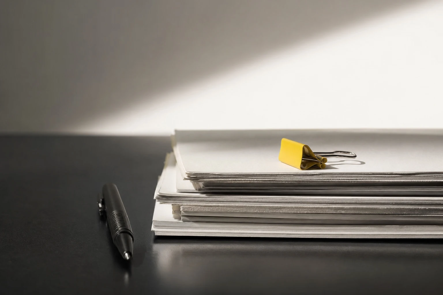Stack of papers topped with a yellow binder clip on a dark desk, with a black pen lying beside it in a shaft of sunlight