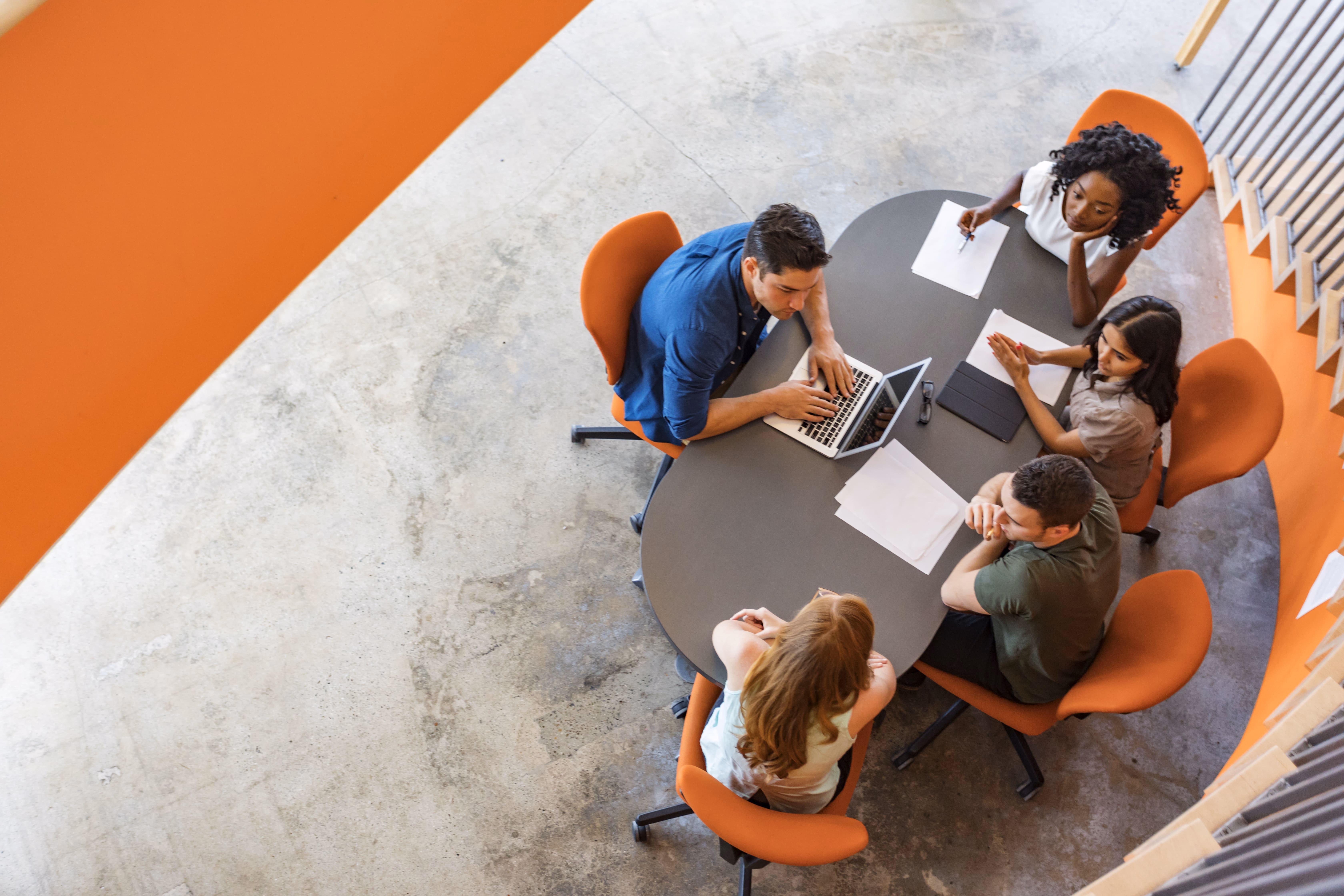 Overhead view of five diverse team members collaborating around a grey circular table with orange chairs