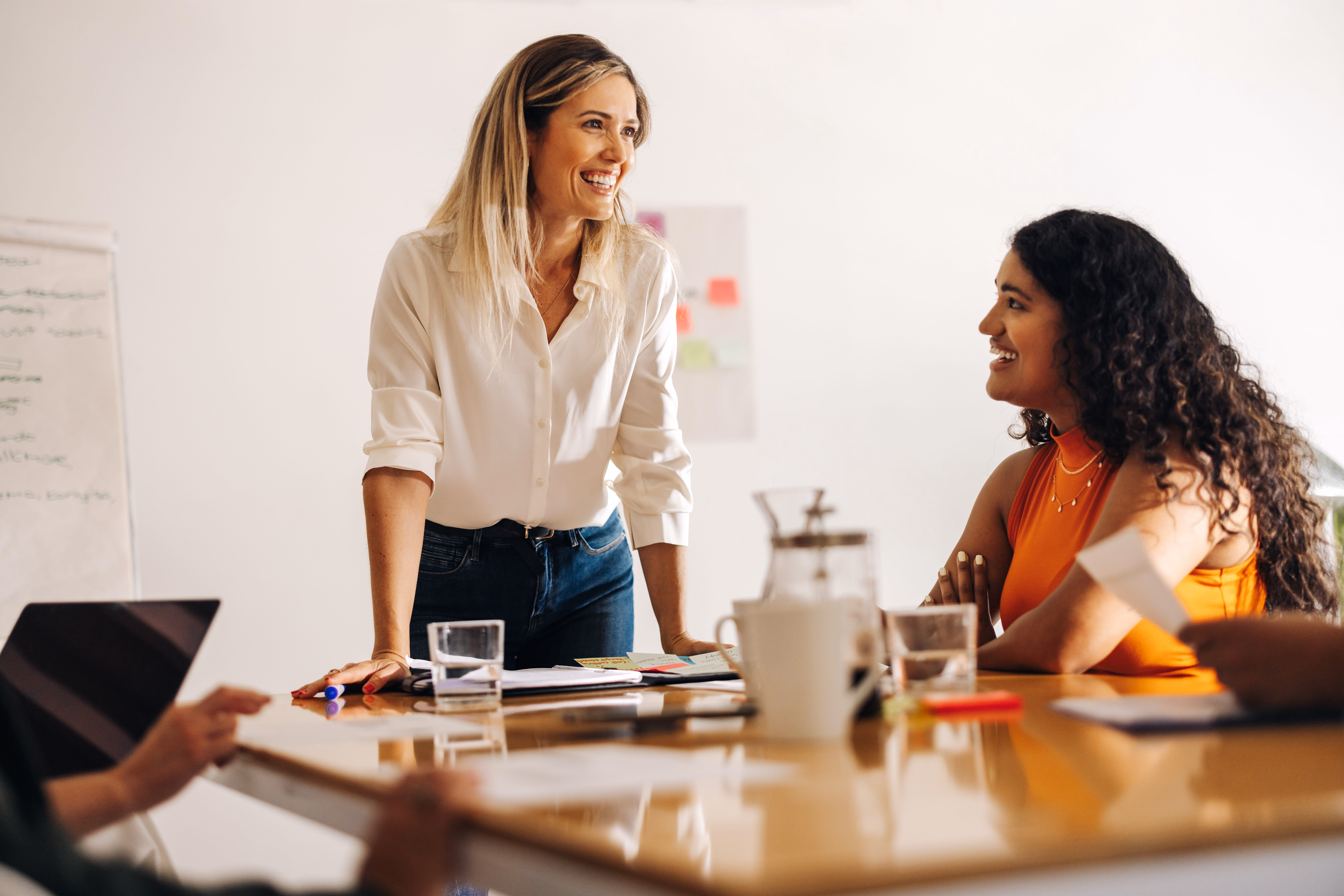Two women collaborating at a meeting table with laptops and planning materials in a bright office