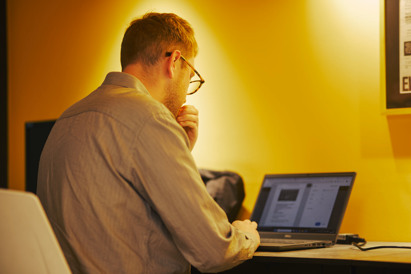 Man wearing glasses works on a laptop at a desk in a warmly lit office