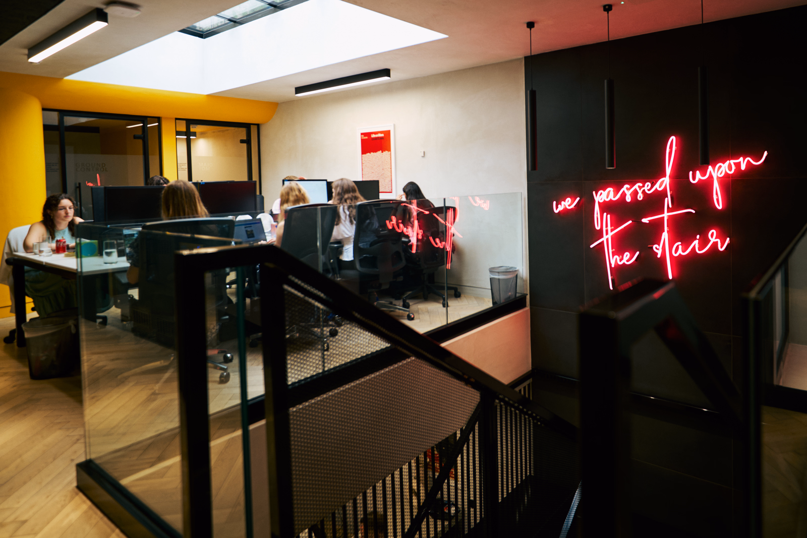 People working at desks in a modern office, with a pink neon sign on a black wall and stairs in the foreground
