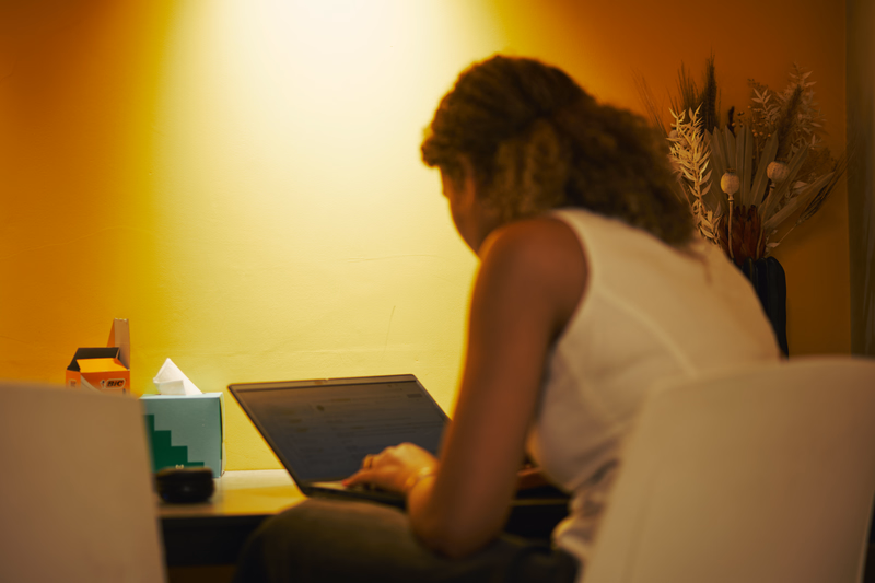 Person working on a laptop at a desk in a warmly lit office, with a tissue box and dried flowers nearby